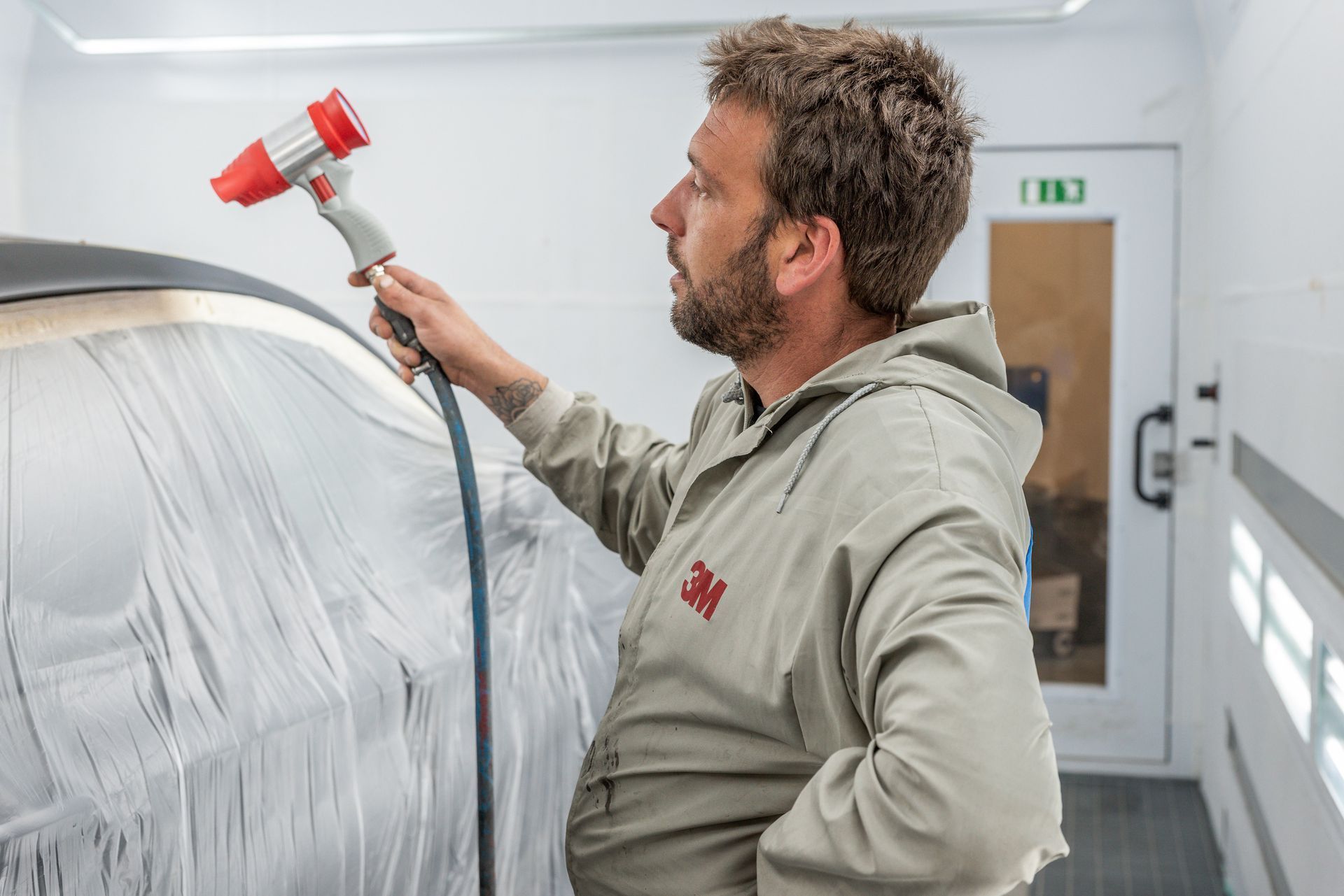 Dans un atelier de carrosserie, un technicien utilise un pistolet à peinture sur une voiture recouverte d'une bâche.