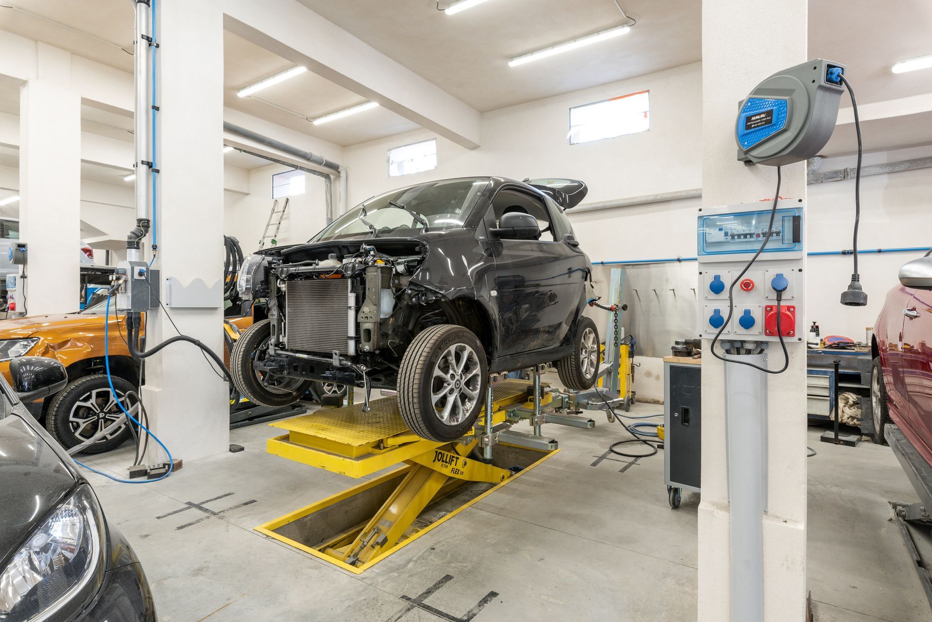 Une voiture noire dont le panneau avant a été démonté est soulevée sur un pont élévateur jaune.