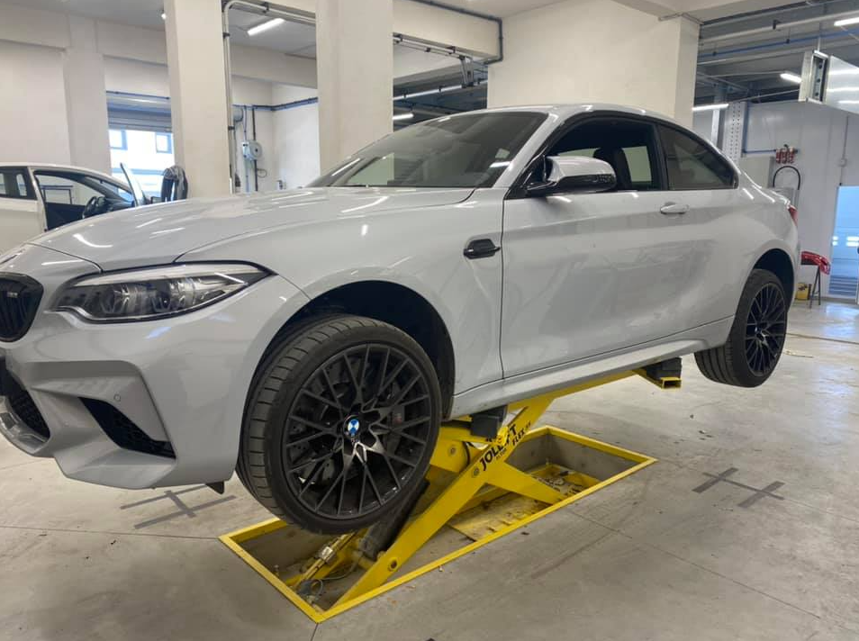 Un coupé BMW M2 gris clair, surélevé sur un pont élévateur jaune.