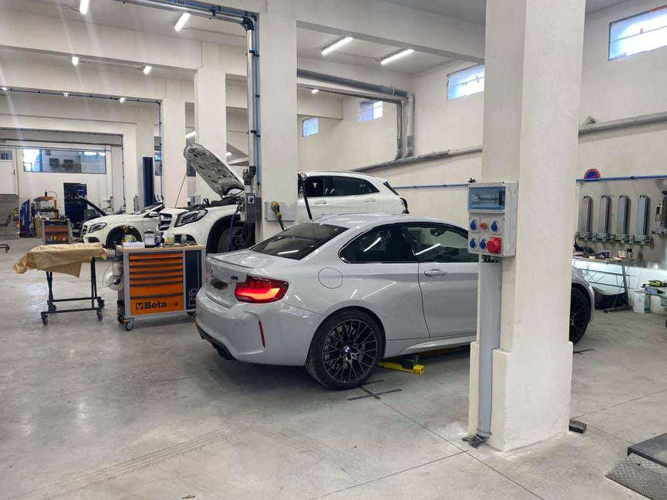 Un garage automobile avec une voiture de sport grise au premier plan et un véhicule blanc, capot ouvert.