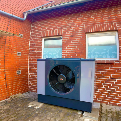 Außenwärmepumpenanlage neben einem roten Backsteinhaus mit zwei Fenstern und gepflastertem Boden