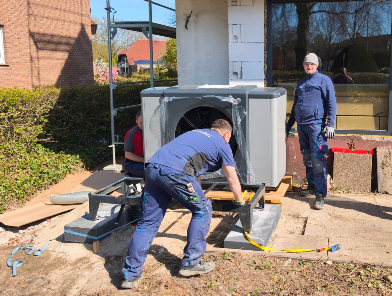 Arbeiter installieren im Freien neben einem Backsteingebäude und einer Betonsäule eine große graue Metallkonstruktion.