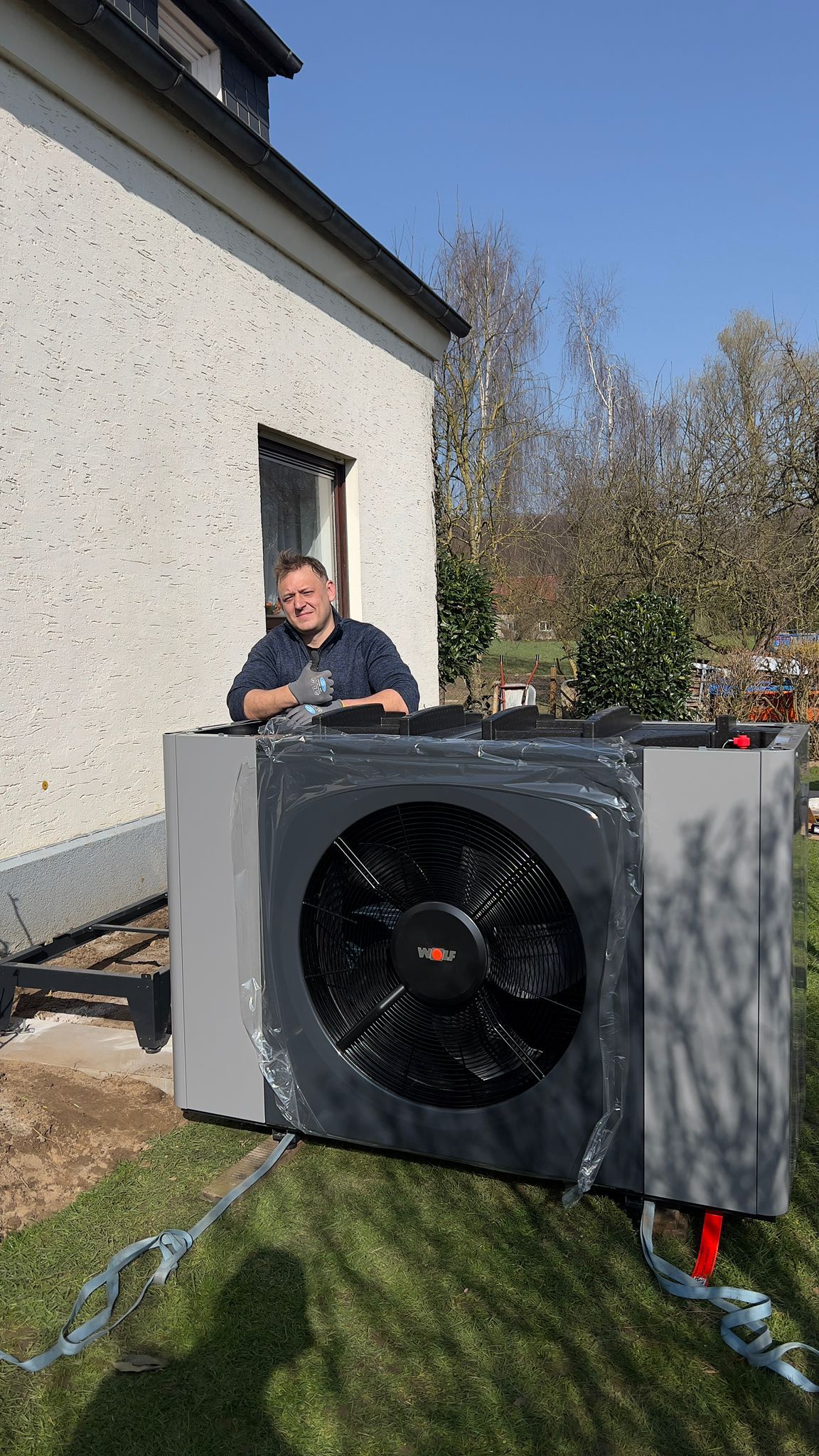 Eine große Außenklimaanlage steht auf einer Rasenfläche neben einem Haus, daneben lehnt eine Person oben drauf.