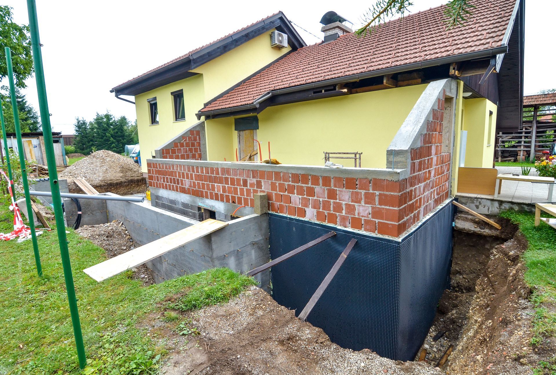 Maison partiellement construite avec mur en briques apparentes, fondations noires et supports en béton dans une cour boueuse.