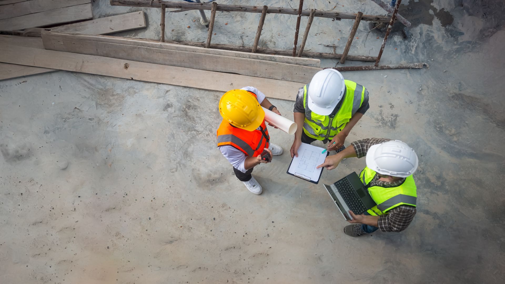 Trois ouvriers du bâtiment, casqués et vêtus de gilets de sécurité, examinent des plans sur un chantier.