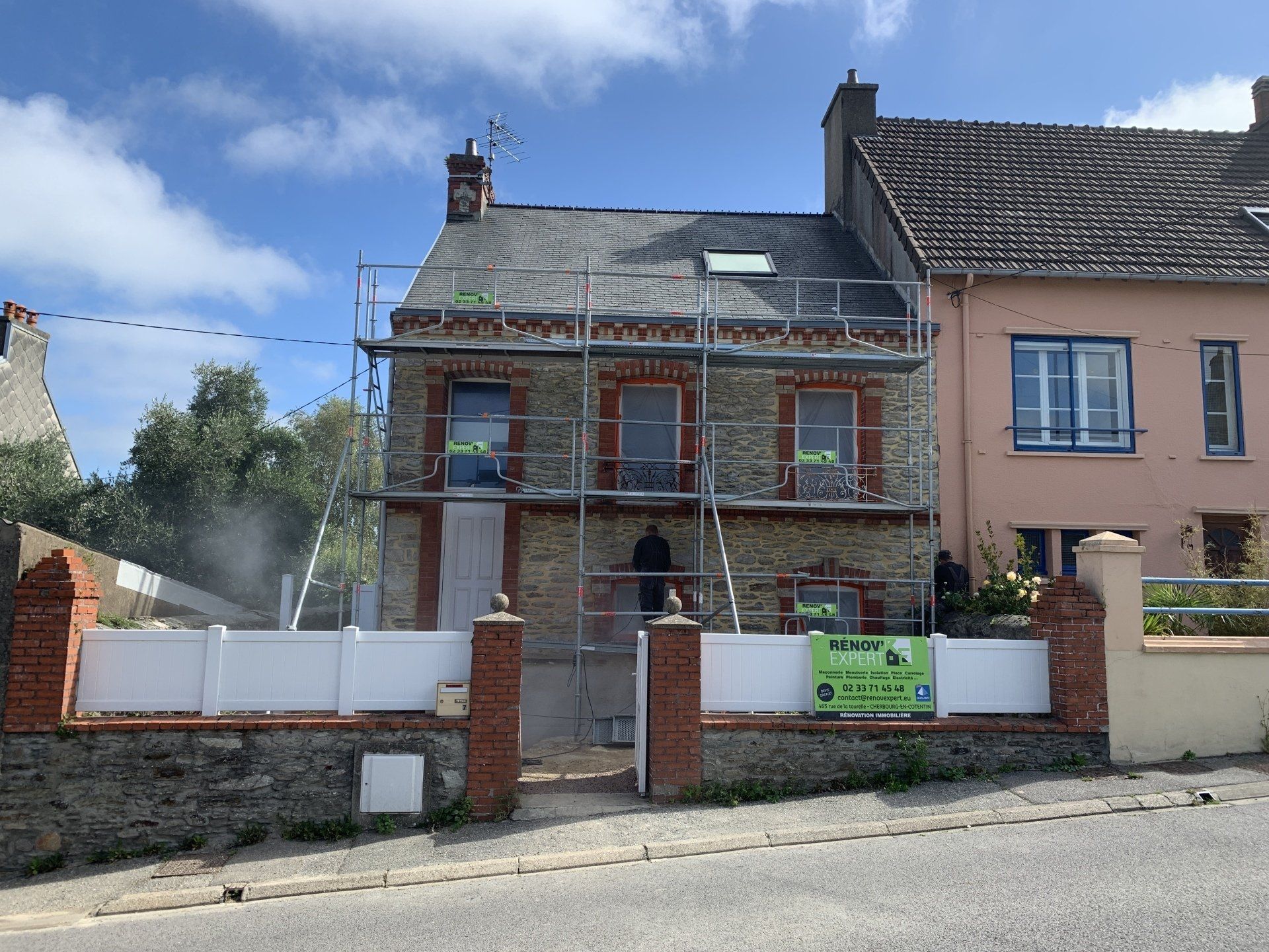Maison partiellement rénovée avec des échafaudages et un panneau vert derrière une clôture en briques blanches.
