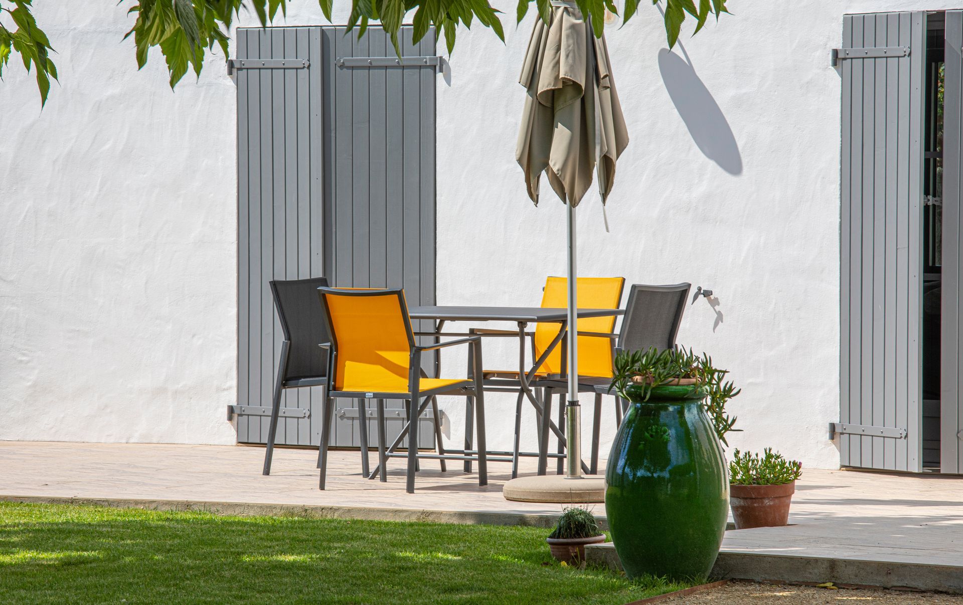 Table avec chaises jaunes et noires sur une terrasse, près d'un mur blanc et de plantes en pot au soleil.