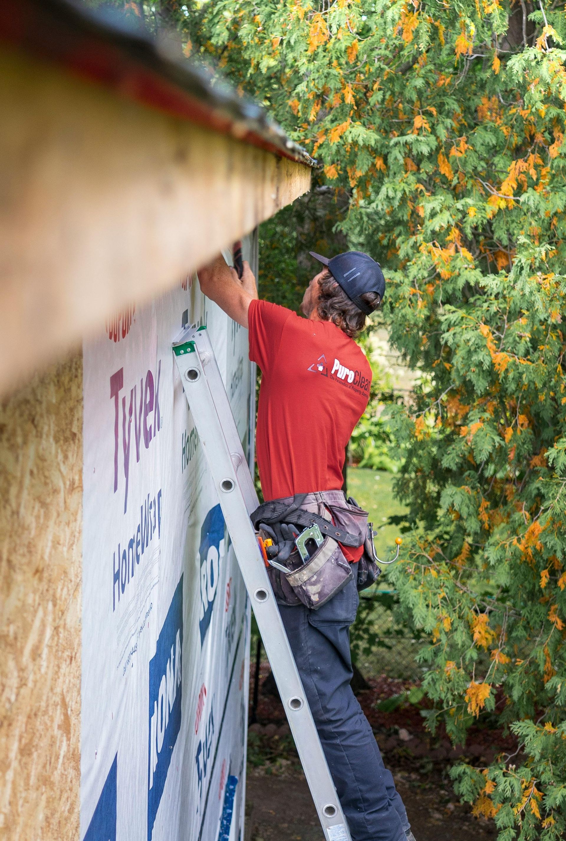 Un ouvrier en chemise rouge pose une toiture ou un bardage sur une maison, à côté d'une échelle et d'arbres.