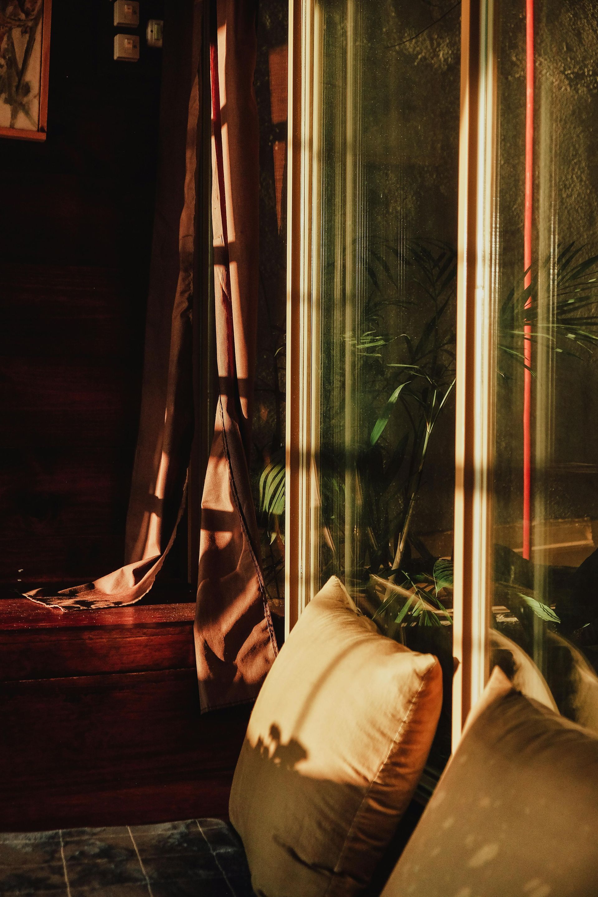 Fenêtre ensoleillée avec voilages, plante en pot et deux coussins beiges près d'un banc en bois.
