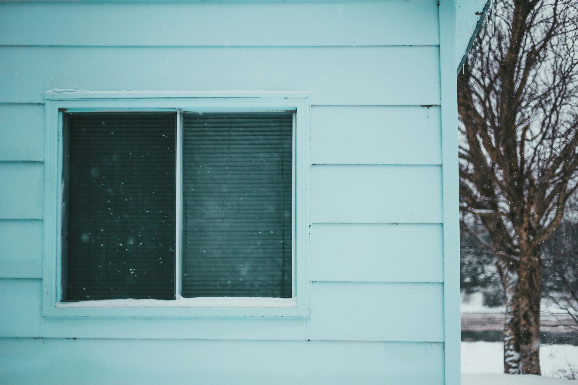 Fenêtre enneigée sur la façade bleu pâle d'une maison, avec des arbres dénudés en arrière-plan.