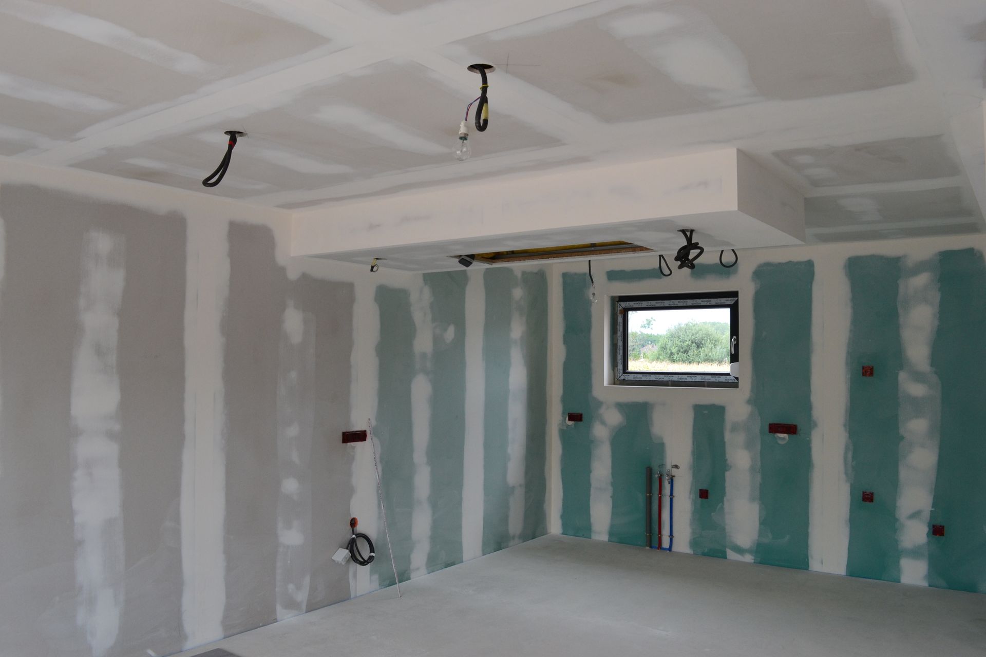 Pièce avec des plaques de plâtre et des joints collés, câblage au plafond et une petite fenêtre sur le mur du fond.