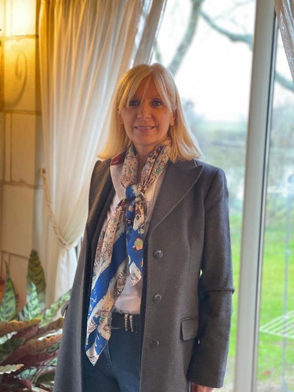 Femme blonde portant un manteau gris et une écharpe à motifs, debout à l'intérieur près d'une fenêtre.