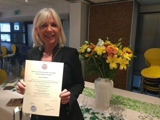 Une femme tenant un certificat, souriante près d'un vase de fleurs.