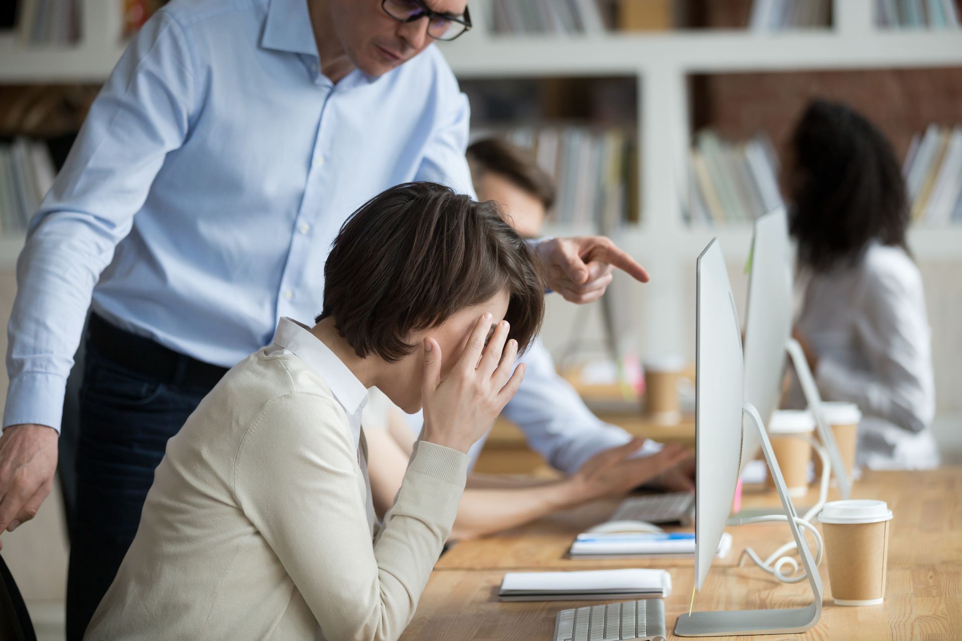 Un homme pointe du doigt un écran d'ordinateur, une femme se prend la tête entre les mains, assise à un bureau, dans un environnement de bureau.