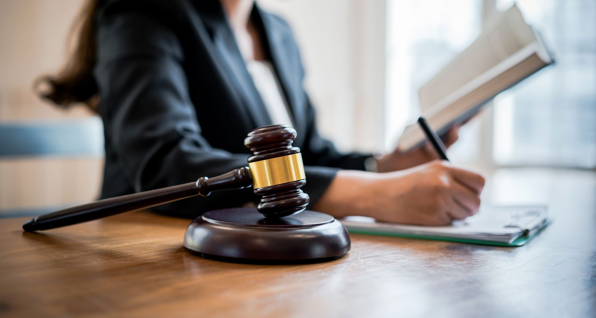 Un maillet est posé sur un bureau au premier plan ; une personne en veste de costume écrit sur un bloc-notes et regarde un livre à l'arrière-plan.