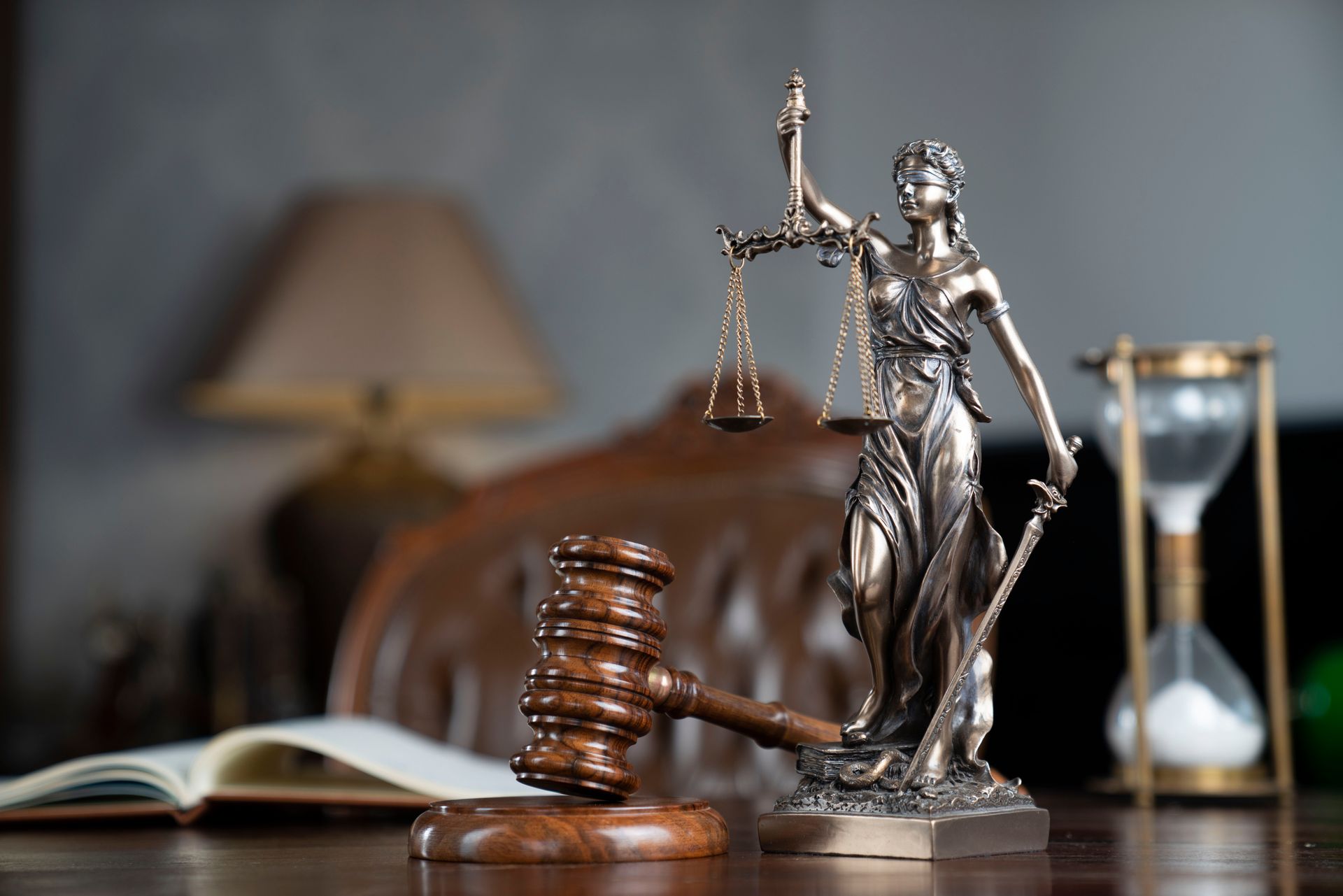 Une statuette de la Justice et un marteau posés sur un bureau, avec un livre ouvert et un sablier en arrière-plan.