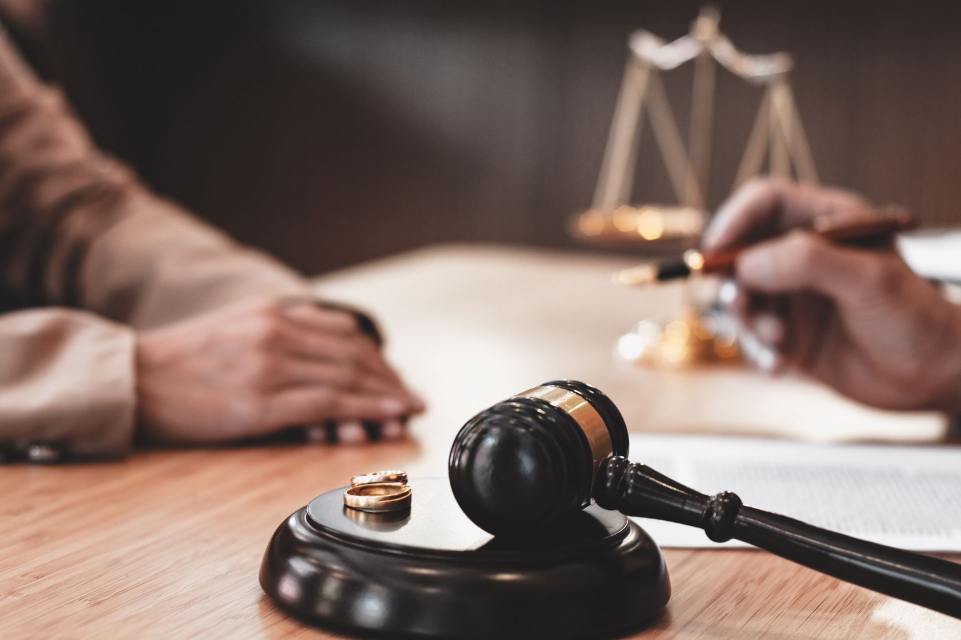 Des alliances à côté d'un maillet et d'une balance de la justice sur une table en bois, avec des mains en arrière-plan.