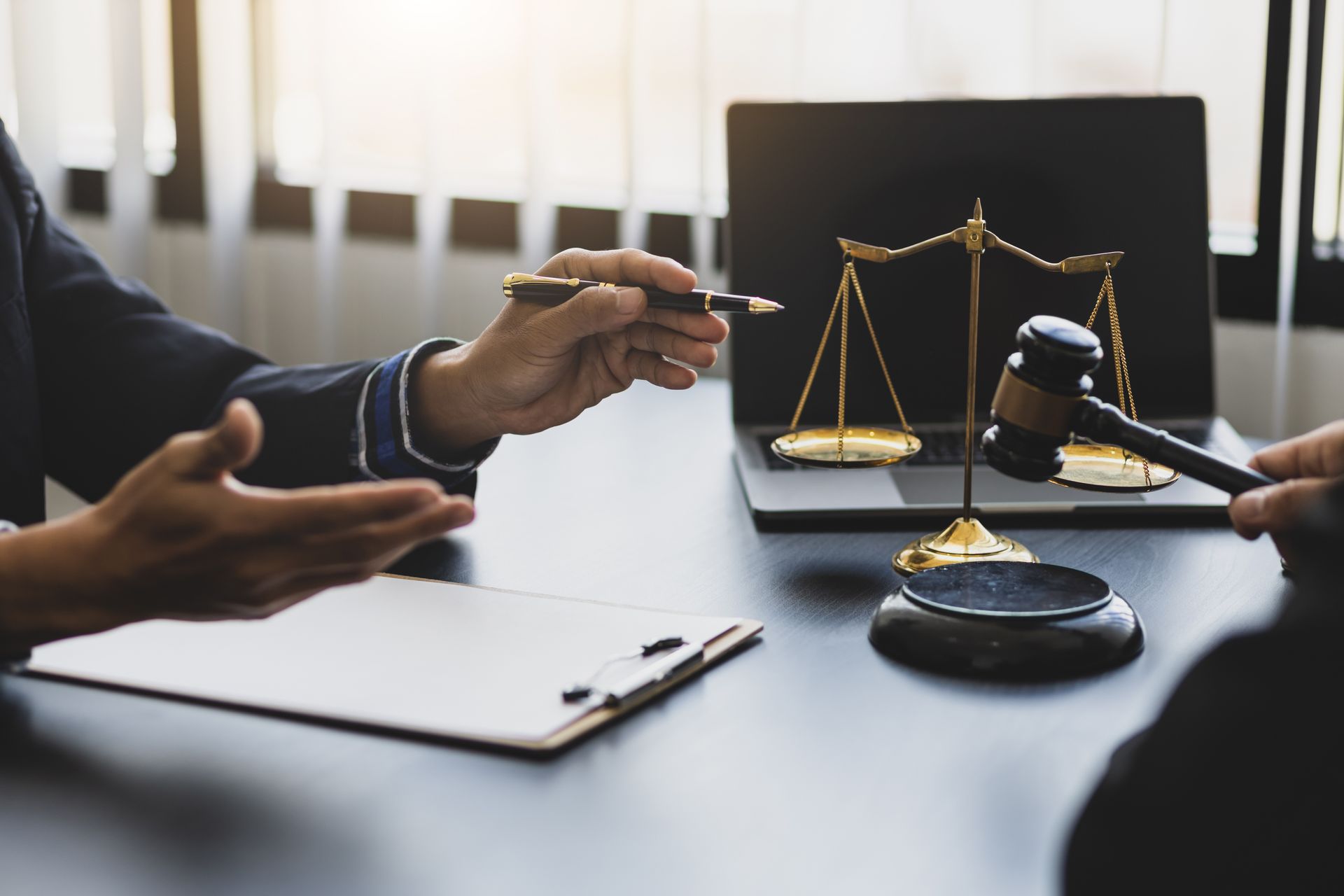 Deux personnes assises à un bureau ; l'une pointe un stylo, l'autre tient un maillet près d'un ordinateur portable et d'une balance de la justice.
