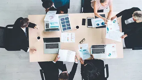 Un grupo de personas están sentadas alrededor de una mesa con computadoras portátiles y papeles.