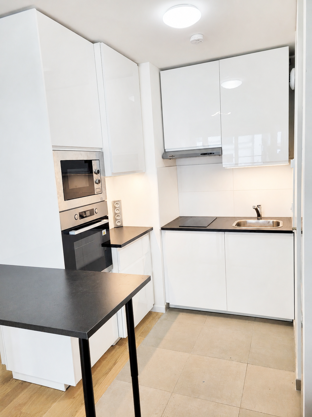 Cuisine moderne blanche avec comptoirs noirs, four encastré, évier et petite table, vue depuis la porte