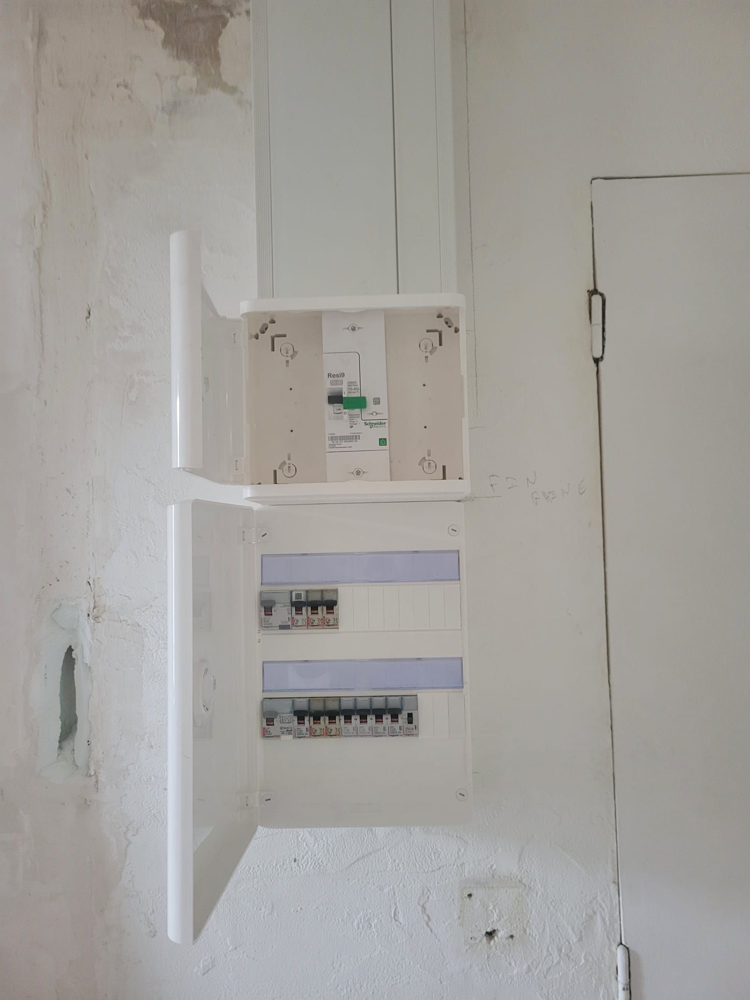 Tableau électrique ouvert sur un mur blanc, avec fils et disjoncteurs apparents.