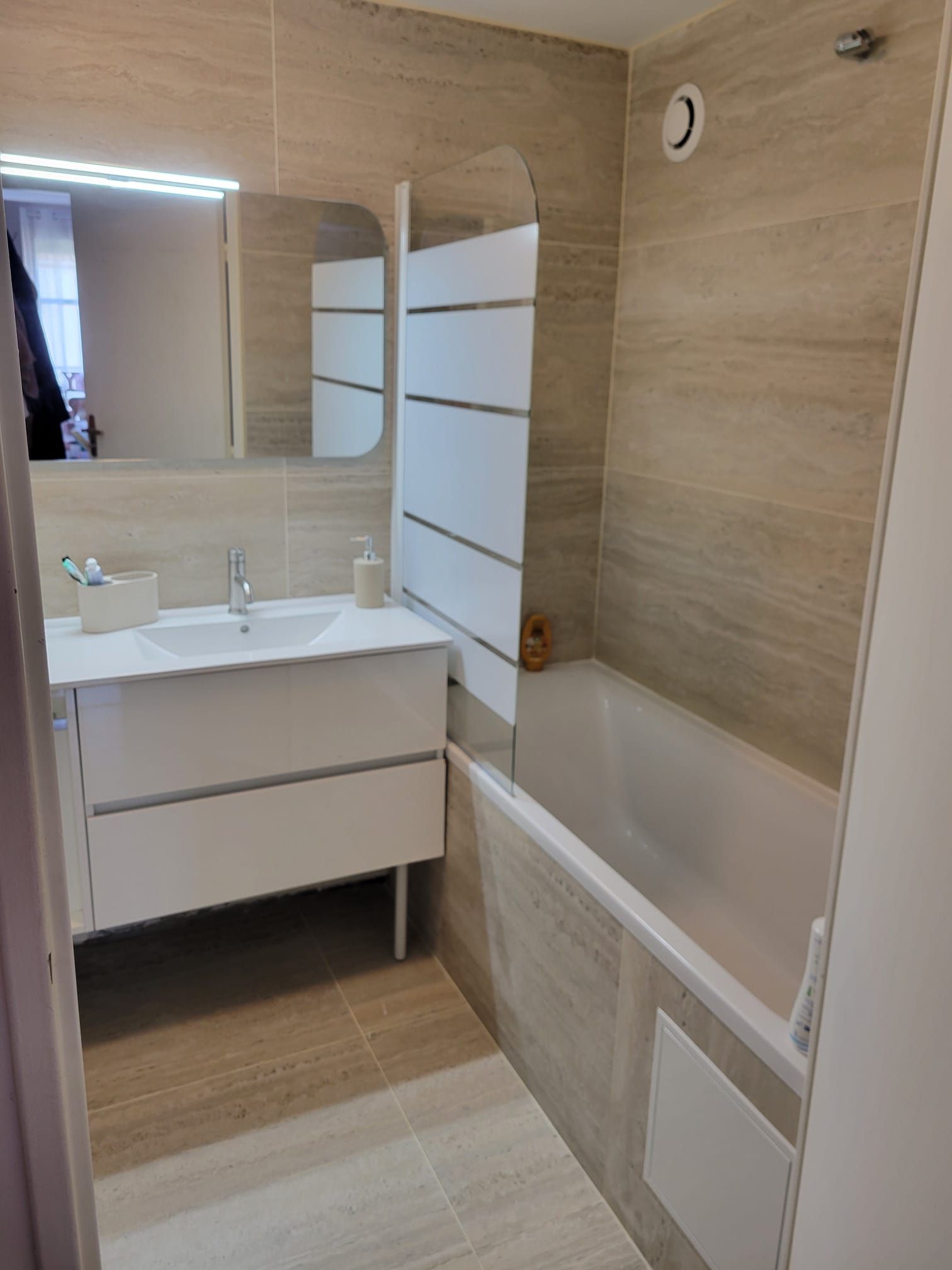 Salle de bains moderne avec meuble-lavabo blanc, miroir, murs carrelés beiges et baignoire avec paroi de verre.