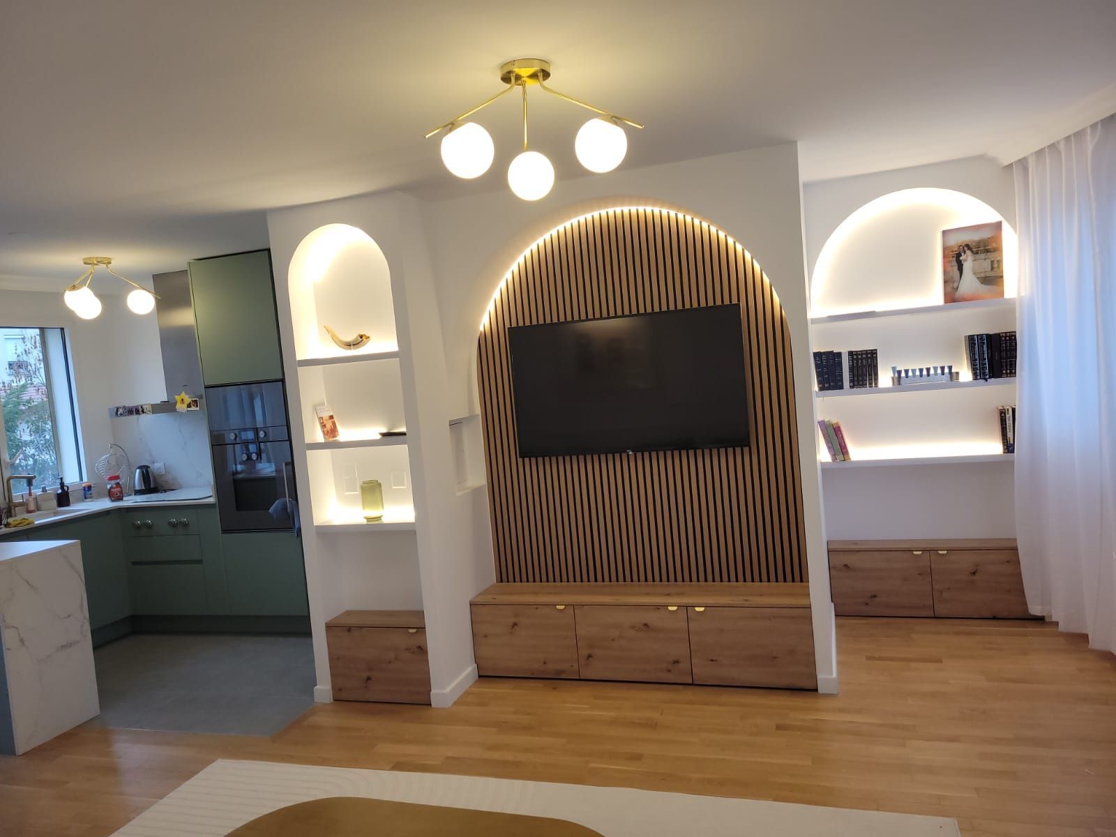 Salon lumineux avec coin TV intégré, étagères cintrées, parquet et vue ouverte sur la cuisine.