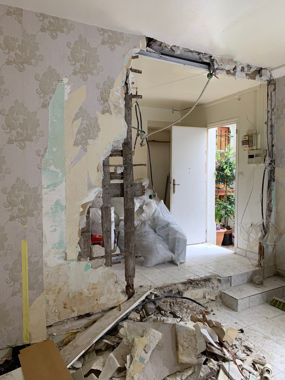 Mur intérieur endommagé, jonché de gravats et de débris, révélant une porte et une pièce lumineuse au-delà.