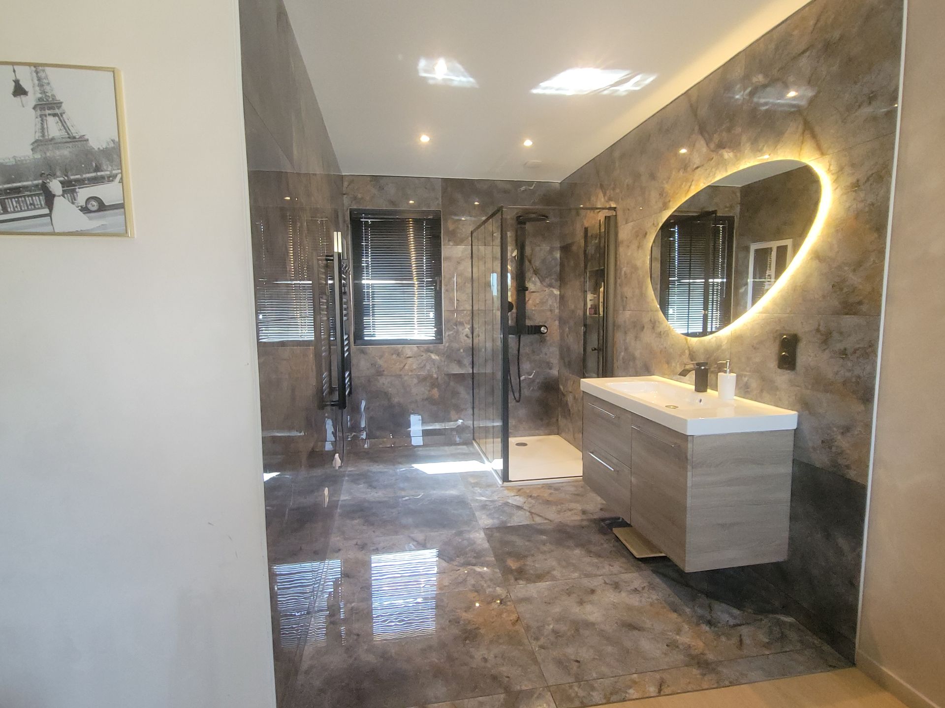 Salle de bains moderne avec murs en marbre gris, douche en verre, meuble-lavabo blanc et miroir ovale éclairé.