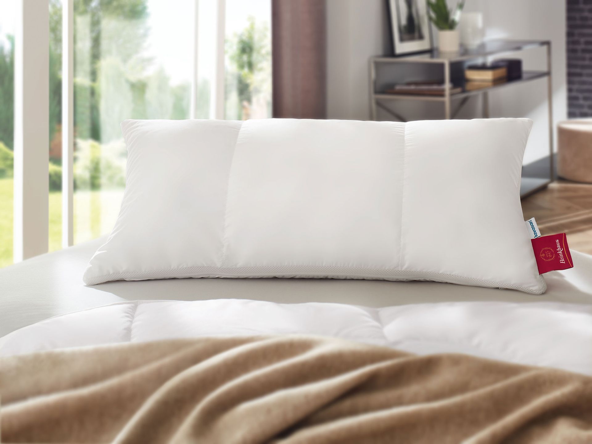 Weißes Kissen auf einem Bett mit beiger Decke. Innenaufnahme mit einem Fenster im Hintergrund.