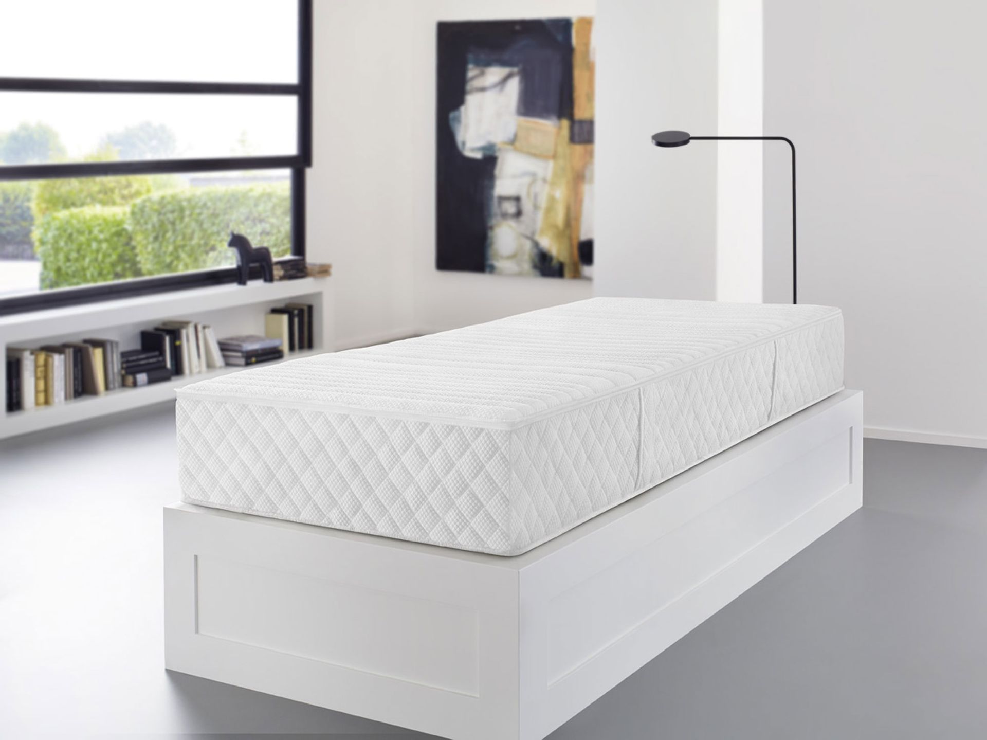 Weiße Matratze auf einem weißen Bettgestell in einem modernen Zimmer mit Kunstwerken und einem Fenster.