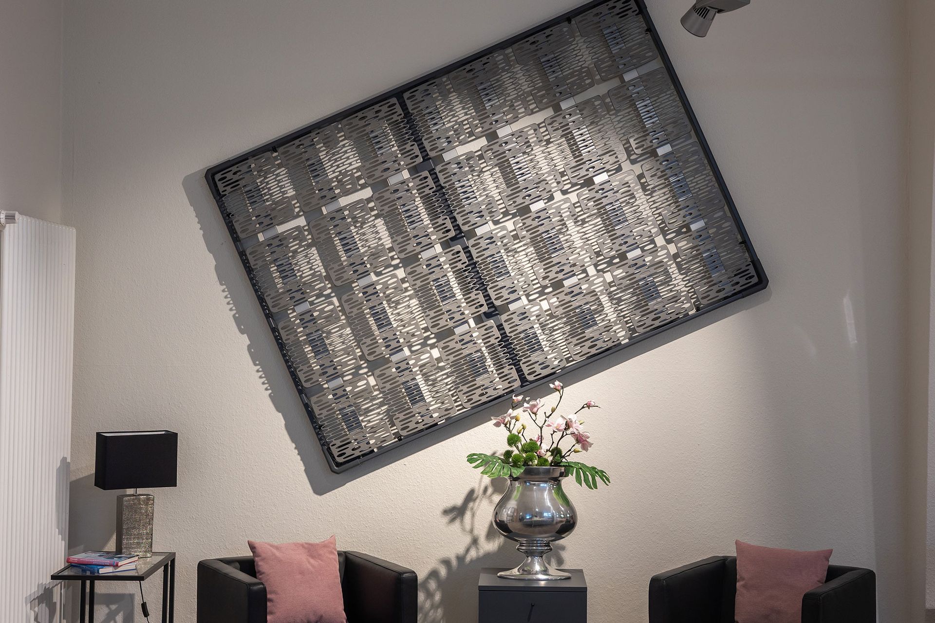 Moderne Kunstinstallation an einer Wand, davor schwarze Sessel und Blumen in einer Vase.