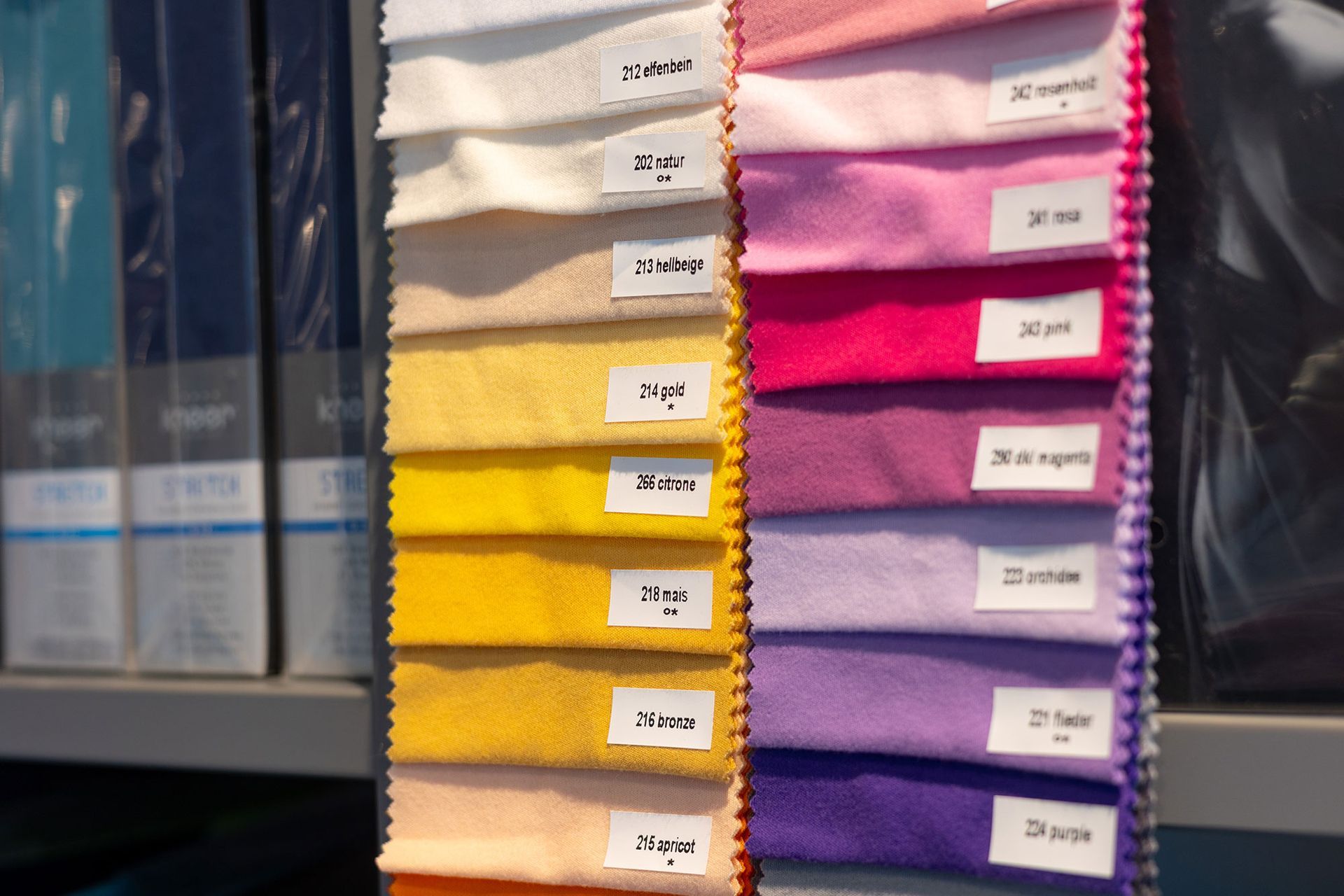 Stoffmuster in verschiedenen Farben, mit Nummern und Namen versehen, auf einem Gestell ausgestellt.