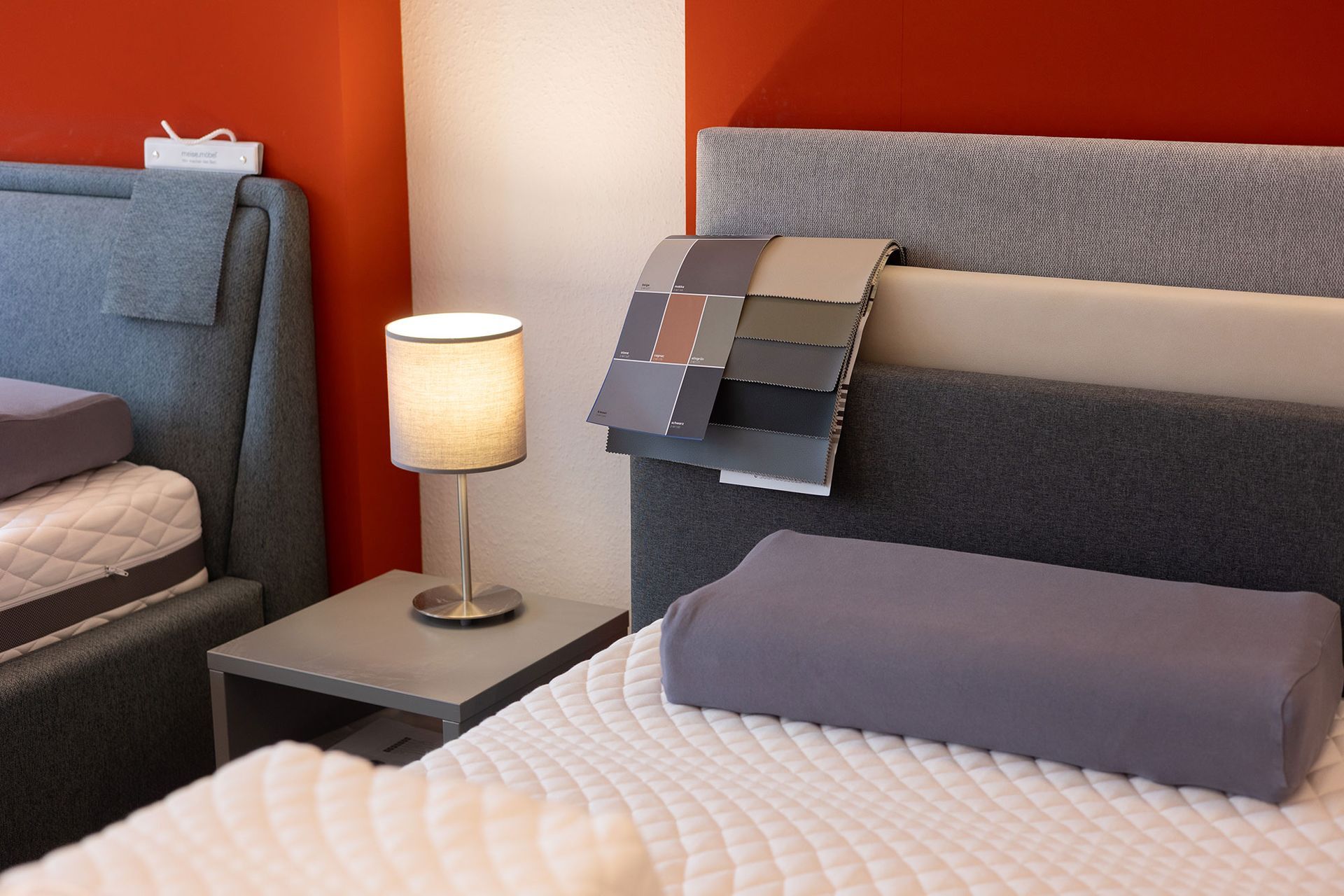 Bettpräsentation mit Kissen, Lampe und Stoffmustern vor rot-weißen Wänden.