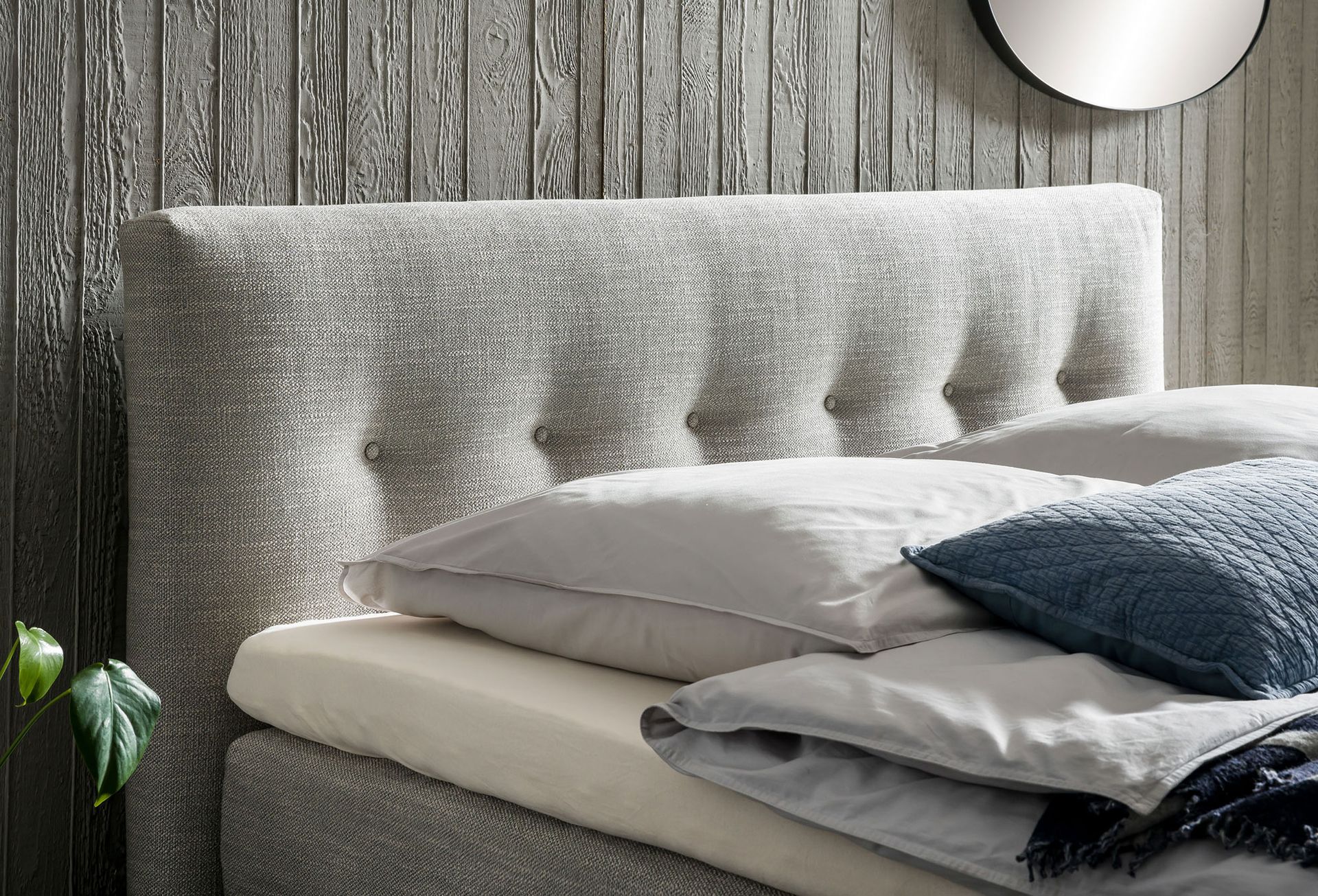 Gepolstertes Bett mit knopfgeheftetem Kopfteil; Kissen und Bettdecke liegen auf dem Bett; dahinter eine Holzwand.