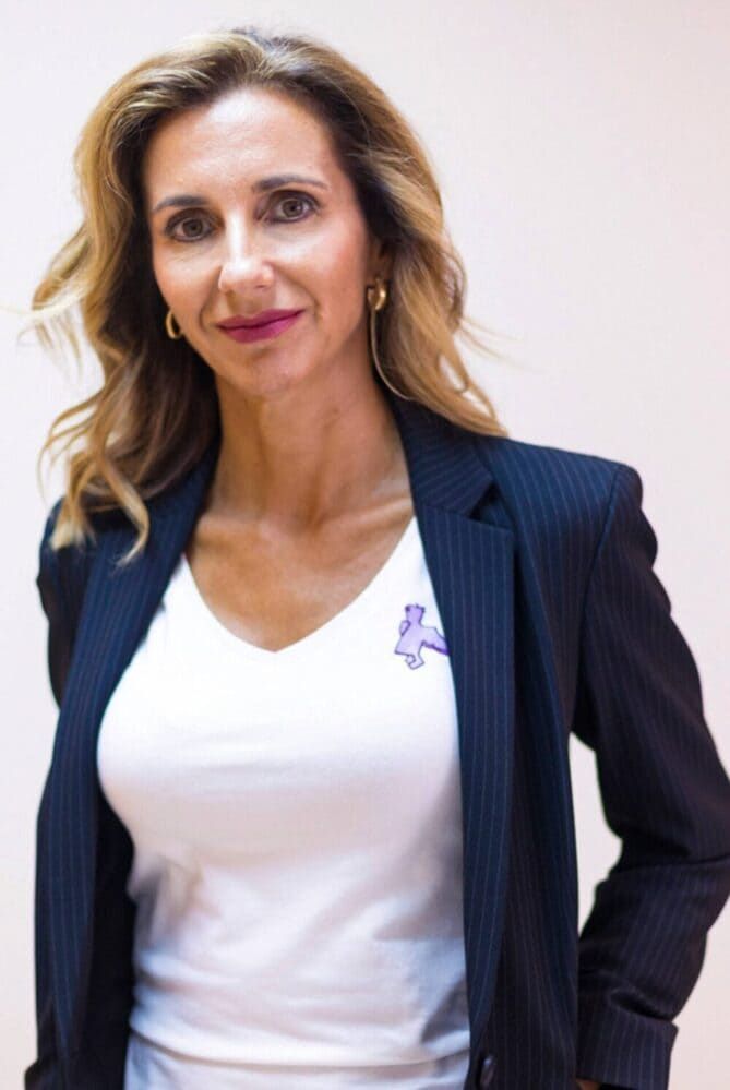 Mujer con una chaqueta azul marino y camisa blanca, de pie frente a un fondo liso y claro.