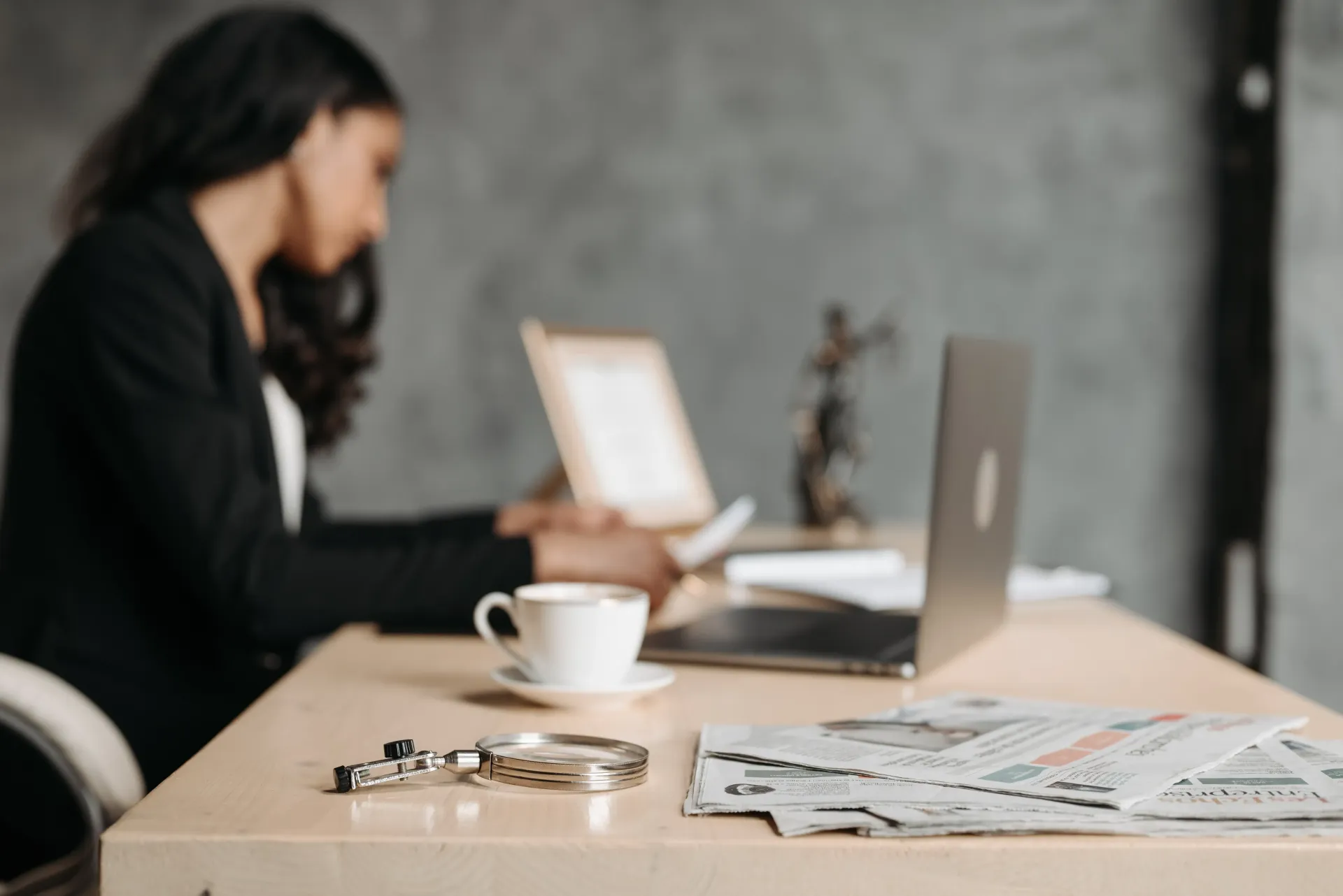 Mujer en un escritorio con un ordenador portátil y un café; esposas y periódico en primer plano.