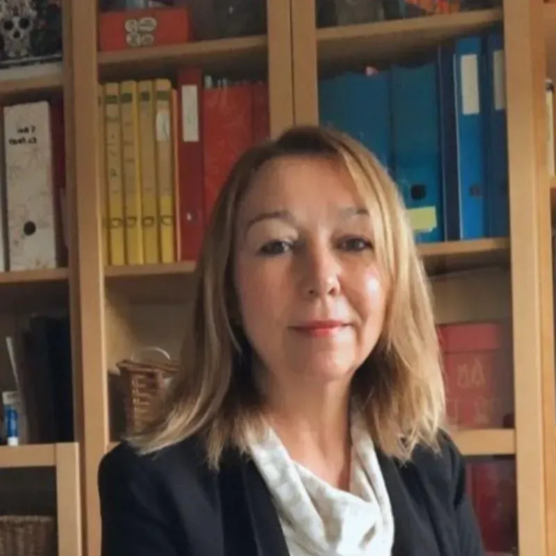 Mujer con cabello rubio hasta los hombros, vistiendo un blazer negro, mirando a la cámara, estanterías en el fondo.