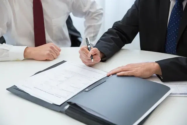 Dos hombres de traje en una mesa, uno firmando un documento, el otro mirando.