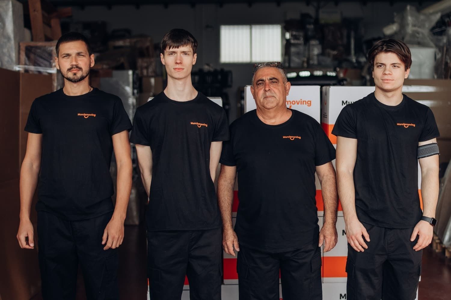 Cuatro hombres con camisas negras de pie en un almacén, con cajas detrás de ellos.