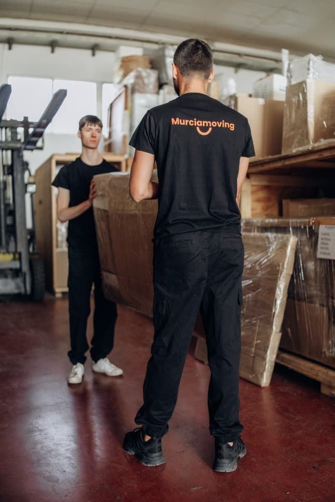Dos trabajadores de mudanzas en un almacén cargando un objeto grande envuelto. Uno lleva una camiseta que dice 