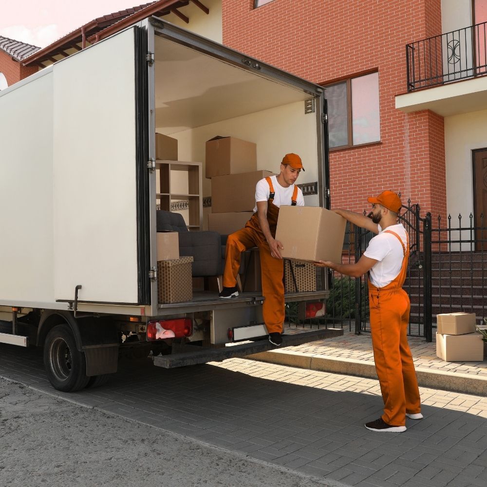 Dos operarios de mudanzas cargan una caja en un camión estacionado frente a una casa.
