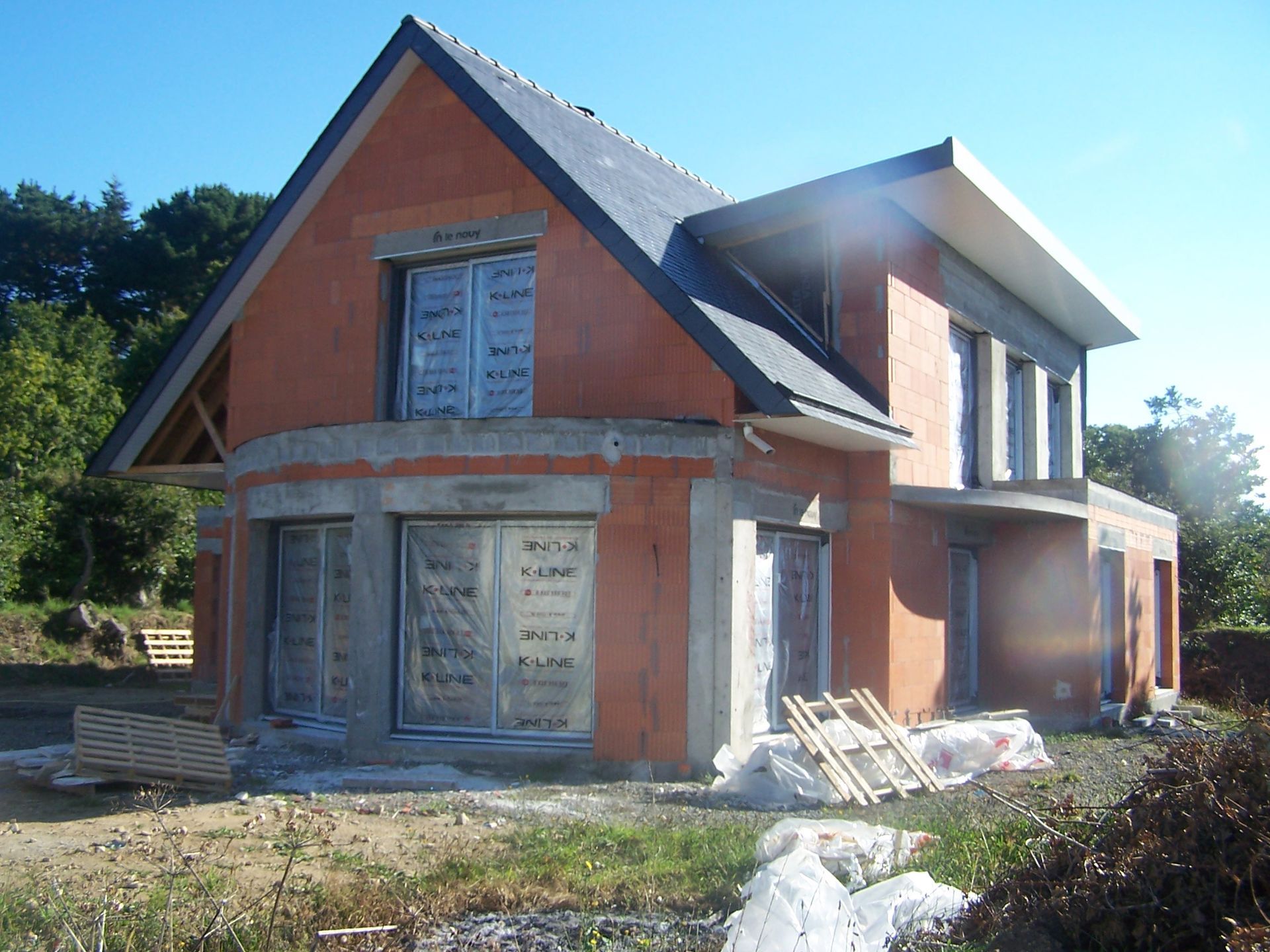 Maison à deux étages partiellement construite, avec des murs en briques et un toit gris, dans une cour ensoleillée.