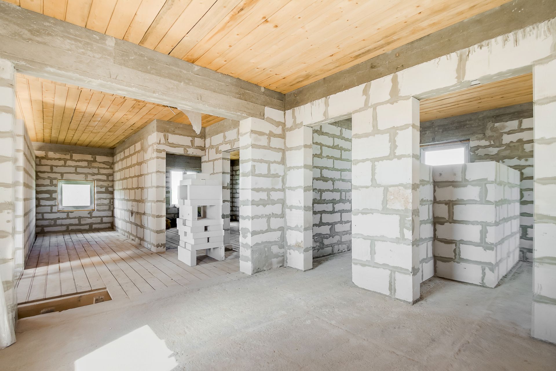 Intérieur d'une maison en parpaings inachevée, avec murs en béton apparent, plafond en bois et portes ouvertes.