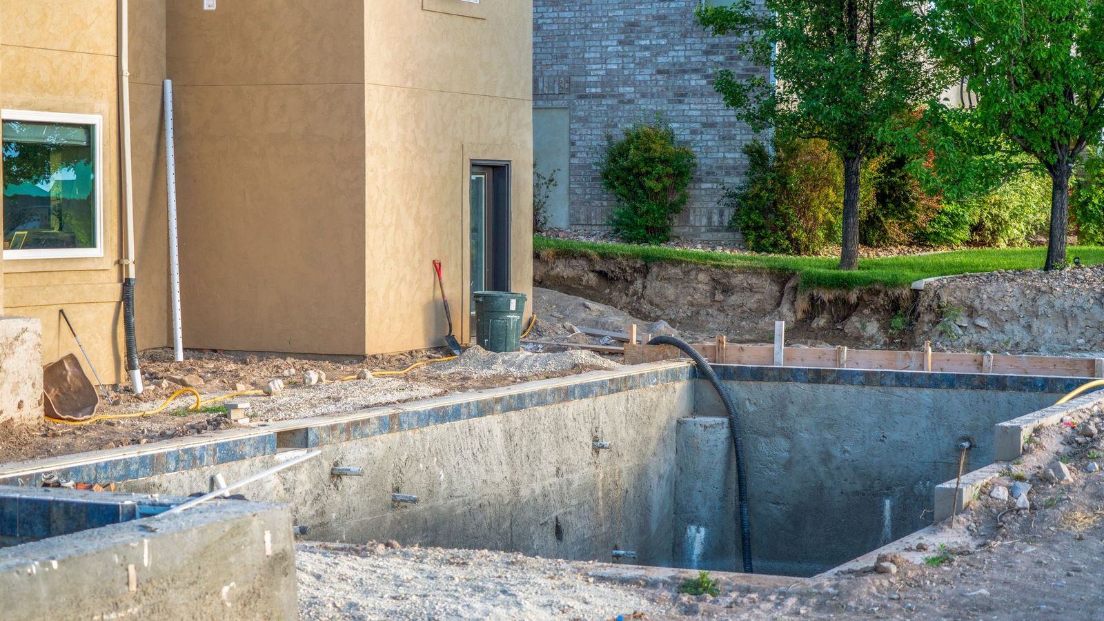 Piscine partiellement construite dans une cour arrière, avec murs en béton, jouxtant une maison beige et un muret paysager en pierre.