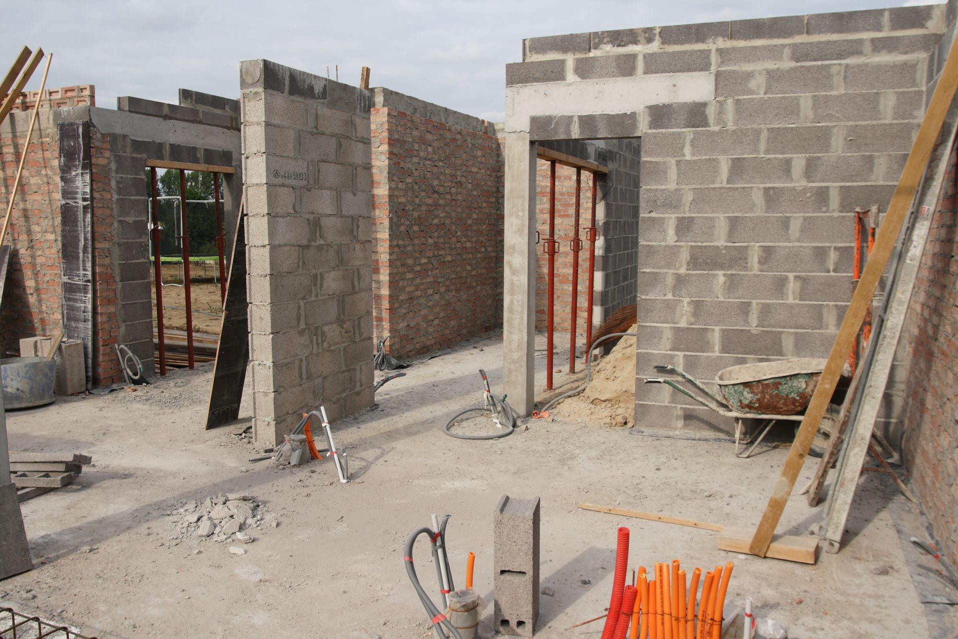 Intérieur d'une maison en parpaings partiellement construite, avec portes ouvertes, matériaux de construction et barres d'armature apparentes.