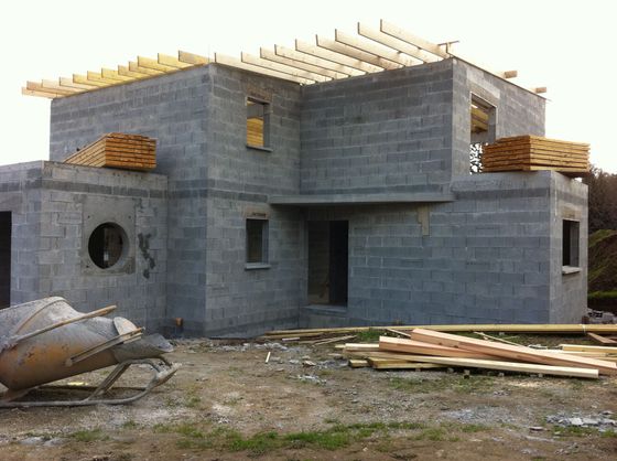 Maison en parpaings gris partiellement construite, avec poutres de toit, débris de construction et une bétonnière à proximité.