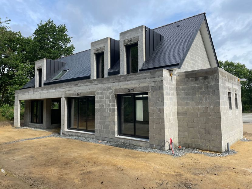 Un ouvrier pose des blocs de béton gris pour un mur. Il porte des gants jaunes et un gilet de sécurité.