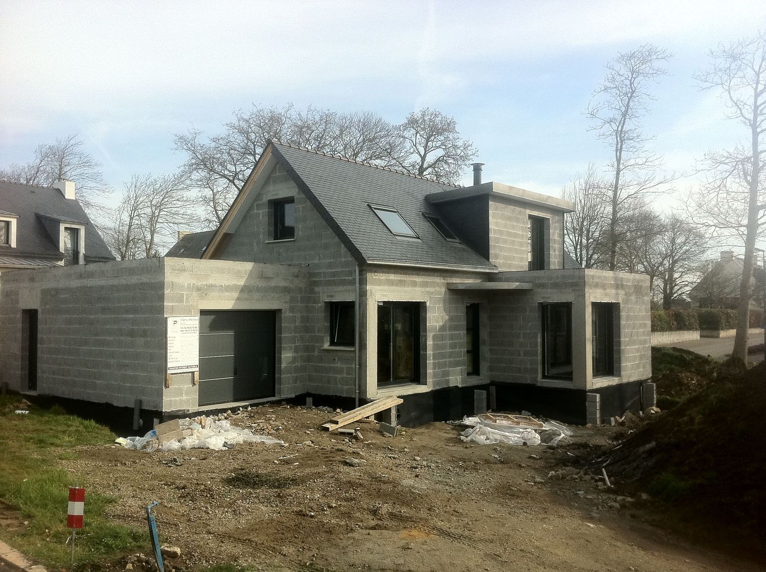 Maison grise partiellement construite, avec bardage et toiture inachevés, et un chantier de terre autour.