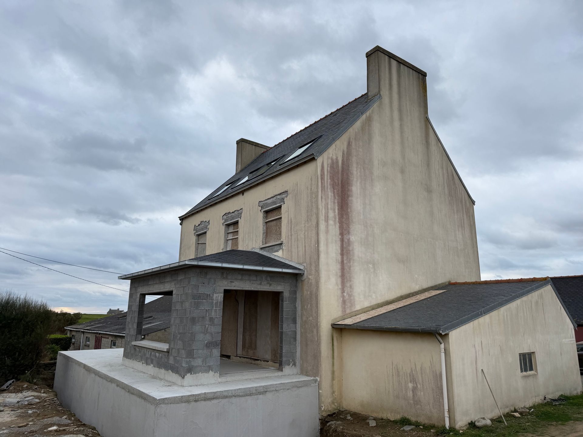 Maison moderne grise avec de grandes fenêtres et un petit patio, se détachant sur un fond de montagnes et de ciel nuageux.